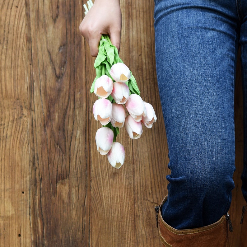 May include: A bouquet of pale pink tulips held by a person wearing blue jeans and brown boots.
