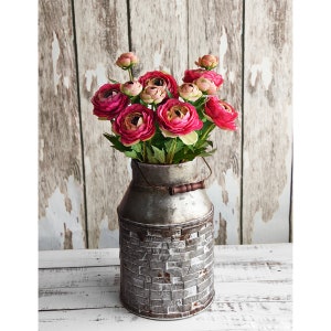 May include: A rustic metal milk can vase filled with vibrant pink and peach flowers. The vase has a brick-like pattern on the bottom and a wooden handle. The background is a distressed white and brown wooden wall.