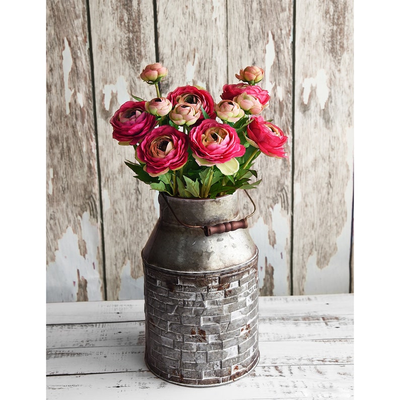 May include: A rustic metal milk can vase filled with vibrant pink and peach flowers. The vase has a brick-like pattern on the bottom and a wooden handle. The background is a distressed white and brown wooden wall.