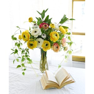 May include: A vibrant floral arrangement in a clear glass vase, featuring yellow, white, and pink ranunculus flowers, accented with green foliage. An open book rests on a white tablecloth, creating a serene, inviting scene.