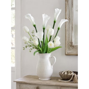 May include: A white ceramic pitcher vase filled with an arrangement of white calla lilies and white tulips. The flowers feature green stems and leaves, accented by small sprigs of green foliage. The pitcher has a handle and a distressed finish.