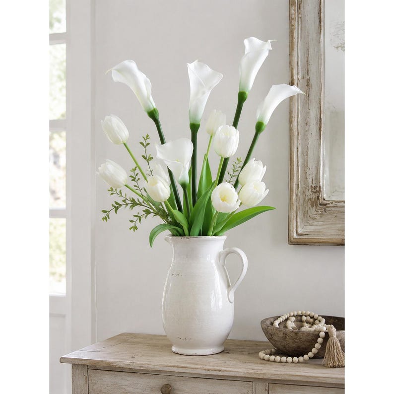 May include: A white ceramic pitcher vase filled with an arrangement of white calla lilies and white tulips. The flowers feature green stems and leaves, accented by small sprigs of green foliage. The pitcher has a handle and a distressed finish.