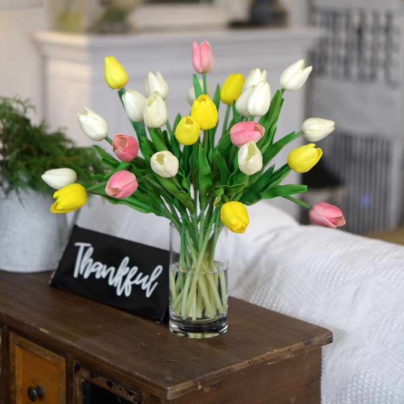 K&ouml;nnte beinhalten: Ein Strau&szlig; k&uuml;nstlicher Tulpen in einer durchsichtigen Glasvase. Die Tulpen sind gelb, wei&szlig; und rosa. Die Vase steht auf einem Holztisch mit einem schwarzen Schild, auf dem "Thankful" steht.