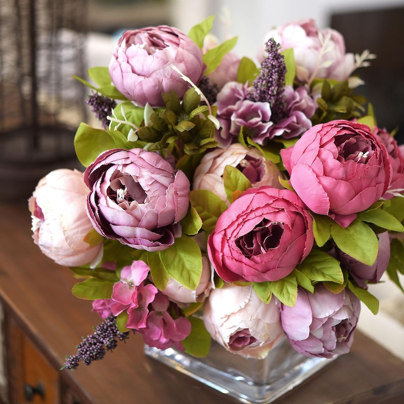 Puede incluir: Un ramo de peon&iacute;as artificiales de color rosa en un jarr&oacute;n de vidrio transparente. Las flores est&aacute;n dispuestas en una variedad de tonos de rosa, desde rosa claro hasta rosa oscuro.