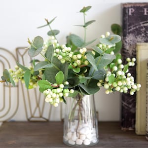 Może przedstawiać: Bukiet sztucznych gałązek eukaliptusa z białymi jagodami w przezroczystym szklanym wazonie wypełnionym białymi kamieniami.