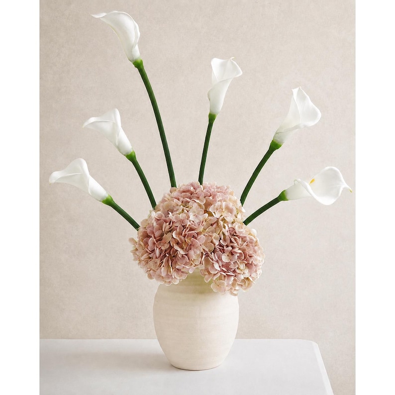May include: A floral arrangement in a cream-colored ceramic vase featuring a cluster of dusty pink hydrangea blossoms at the base and six long-stemmed white calla lilies extending upwards in a fan-like display against a neutral background.