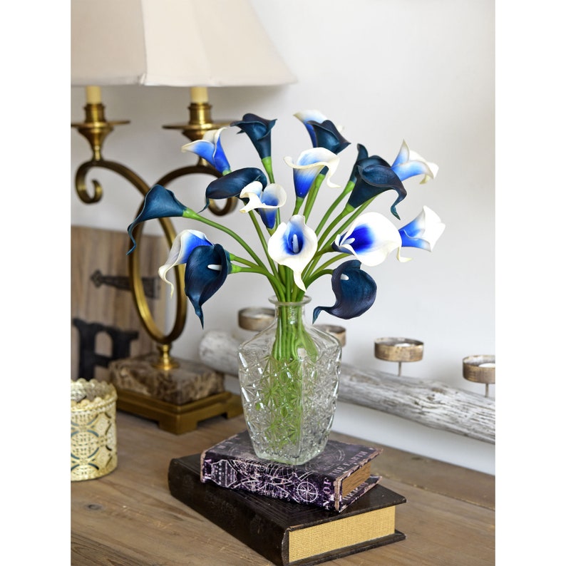 May include: A bouquet of white and blue calla lilies in a clear glass vase with a diamond pattern. The vase is sitting on a stack of books on a wooden table.