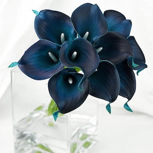 May include: A bouquet of teal blue calla lilies in a clear glass vase. The lilies are arranged in a cluster with the stems facing down.