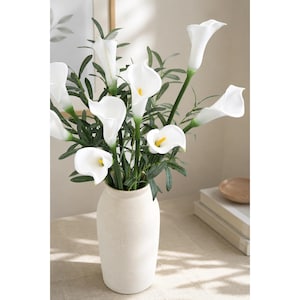 May include: A bouquet of white calla lilies with yellow spadices and green stems and leaves arranged in a tall, matte, off-white ceramic vase. The flowers feature smooth, trumpet-shaped petals and are displayed with slender, pointed green foliage.