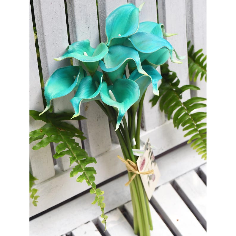 Peut inclure: Un bouquet de callas artificiels d'une couleur turquoise vibrante avec des p&eacute;tales lisses en forme de trompette et des spadices blancs. Les tiges sont regroup&eacute;es par un lien en ficelle naturelle avec une petite &eacute;tiquette en papier rectangulaire.