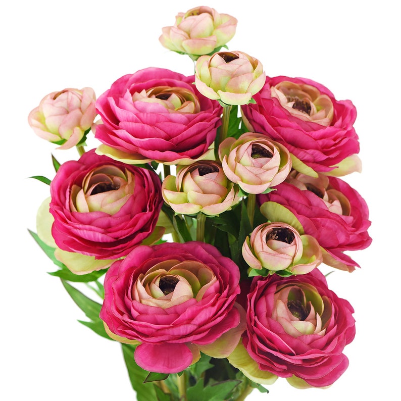 May include: A bouquet of artificial ranunculus flowers in shades of pink and cream. The flowers have multiple layers of petals, with some blooms fully open and others in bud form. Green leaves provide contrast against the white background.
