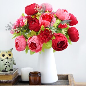 Puede incluir: Un ramo de peonías artificiales rojas y rosas en un jarrón blanco. Las flores tienen hojas verdes y se exhiben en una bandeja de madera con una figura de búho de cerámica, una vela y un tarro.