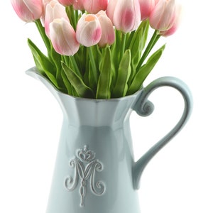 May include: A light blue ceramic pitcher with a decorative design of a crown and the letter "M" on the front. The pitcher is filled with a bouquet of pink artificial tulips.
