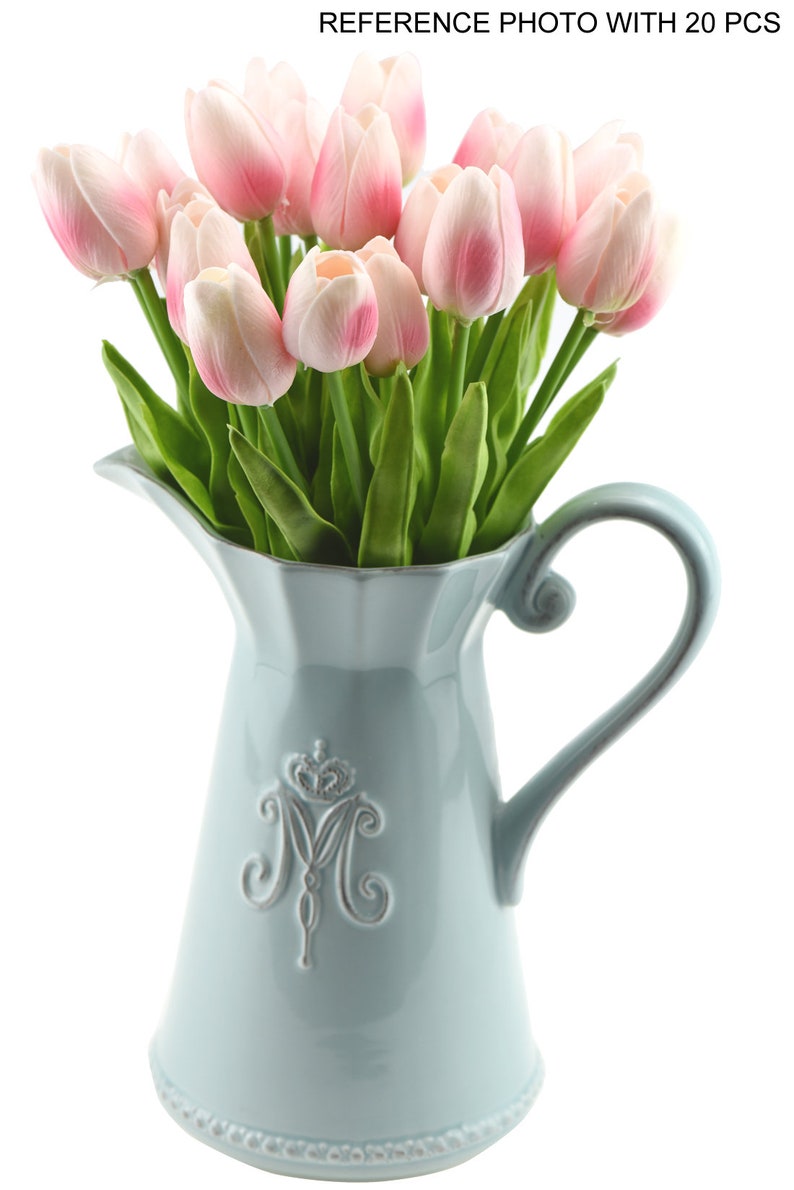 May include: A light blue ceramic pitcher with a decorative design of a crown and the letter "M" on the front. The pitcher is filled with a bouquet of pink artificial tulips.