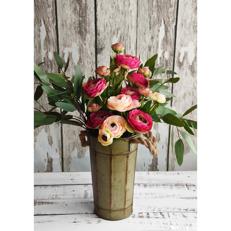 May include: A bouquet of pink and peach flowers with green foliage in a distressed green metal bucket with rope handles. The flowers have multiple layers of petals and are in various stages of bloom.