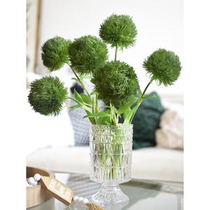 May include: A bouquet of green artificial flowers in a clear glass vase with a geometric pattern. The vase has a pedestal base.