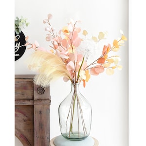 Può includere: Un vaso di vetro trasparente con un'ampia apertura e una superficie testurizzata contiene un bouquet di fiori secchi. Il bouquet comprende rami di eucalipto rosa pallido e giallo, fiori bianchi simili a dente di leone e una grande piuma di erba della pampa di colore crema e soffice.