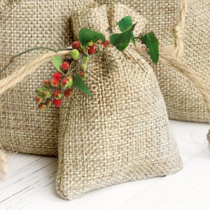 Puede incluir: Dos pequeñas bolsas de arpillera con una rama de bayas rojas y hojas verdes. Las bolsas están atadas con cuerda y están sobre una superficie blanca.