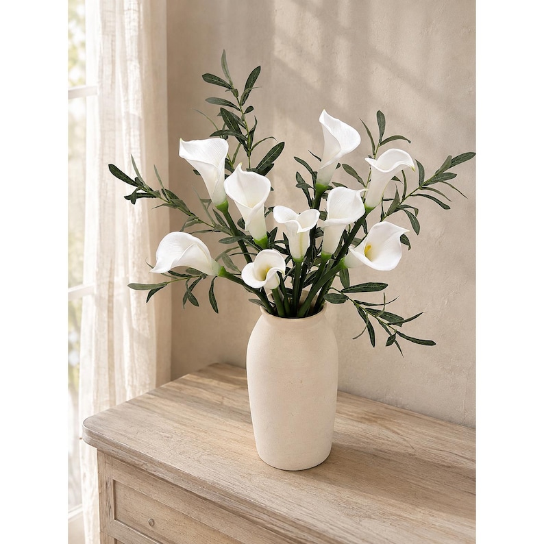 May include: A bouquet of white calla lily flowers with green foliage arranged in a matte, off-white ceramic vase. The flowers feature smooth, trumpet-shaped petals and are interspersed with slender, pointed green leaves, creating a balanced floral display.