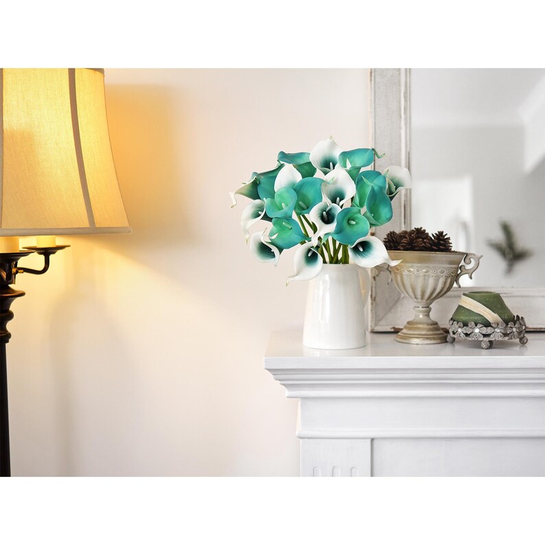 Peut inclure: Un bouquet de callas artificiels pr&eacute;sentant un d&eacute;grad&eacute; de couleurs blanc et sarcelle, dispos&eacute; dans un pichet en c&eacute;ramique blanche. Les fleurs ont des p&eacute;tales lisses et incurv&eacute;s ainsi que des spadices centraux bien visibles.