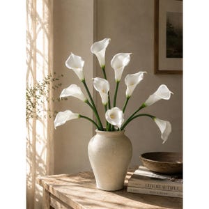 May include: A bouquet of white calla lilies with long green stems arranged in a beige ceramic vase. The flowers feature smooth, trumpet-shaped petals and yellow spadices, creating a balanced floral display with multiple blooms at varying heights.
