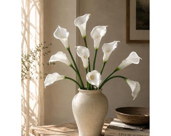 May include: A bouquet of white calla lilies with long green stems arranged in a beige ceramic vase. The flowers feature smooth, trumpet-shaped petals and yellow spadices, creating a balanced floral display with multiple blooms at varying heights.