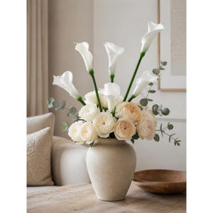 May include: A floral arrangement in a beige ceramic vase featuring white calla lilies, pale peach garden roses, and green eucalyptus stems. The bouquet is dense with layered rose petals and tall, trumpet-shaped lily blooms arranged symmetrically.