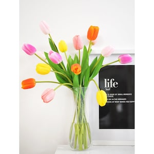 Op de afbeelding: Een boeket kleurrijke kunstmatige tulpen in een heldere glazen vaas. De tulpen zijn roze, geel, oranje en wit. De vaas staat op een wit oppervlak. De tekst "life is made of small moments these joy in the ordinary more of what makes you happy" is gedrukt op een zwarte achtergrond achter de vaas.