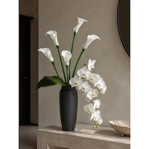 May include: A tall, matte black cylindrical vase containing a floral arrangement of white calla lilies with long green stems and a cascading branch of white phalaenopsis orchids featuring yellow centers and broad green leaves at the base.