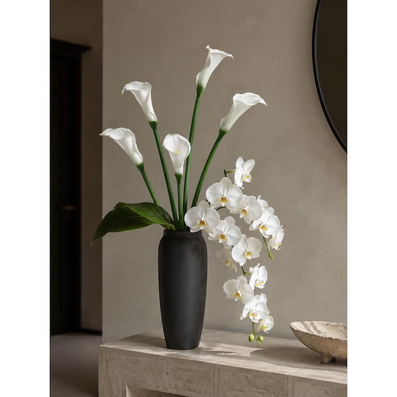 May include: A tall, matte black cylindrical vase containing a floral arrangement of white calla lilies with long green stems and a cascading branch of white phalaenopsis orchids featuring yellow centers and broad green leaves at the base.