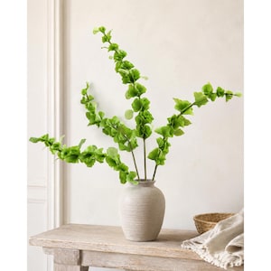 May include: A light beige ceramic vase with a textured, matte finish holds three tall, slender stems of artificial greenery. Each stem features numerous small, ruffled, bright lime-green leaves arranged along the length of the branches.