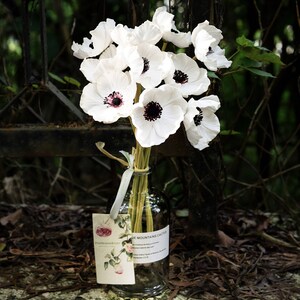 May include: A bouquet of white anemones with black centers arranged in a clear glass vase. The vase is sitting on a bed of brown leaves. A tag with the text "Five Seasons Stuff" and "Blue Mountains Cactus" is attached to the vase.