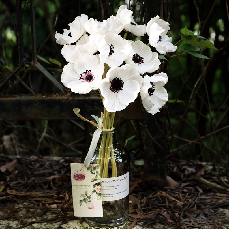 May include: A bouquet of white anemones with black centers arranged in a clear glass vase. The vase is sitting on a bed of brown leaves. A tag with the text "Five Seasons Stuff" and "Blue Mountains Cactus" is attached to the vase.