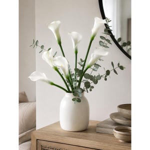 May include: A bouquet of white calla lilies with green eucalyptus stems arranged in a smooth, matte white ceramic vase. The flowers feature long, slender green stalks and trumpet-shaped white petals, creating a clean, balanced floral display.