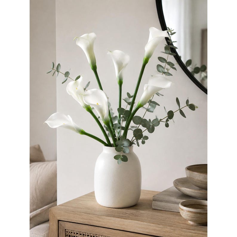 May include: A bouquet of white calla lilies with green eucalyptus stems arranged in a smooth, matte white ceramic vase. The flowers feature long, slender green stalks and trumpet-shaped white petals, creating a clean, balanced floral display.
