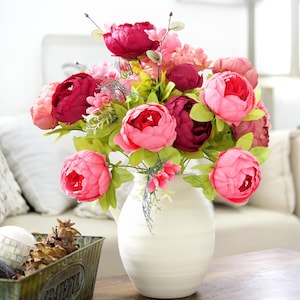 Könnte beinhalten: Ein Strauß aus rosa und burgunderfarbenen Pfingstrosen in einer weißen Keramikvase. Die Blumen haben grüne Blätter und sind in einer üppigen Anordnung angeordnet. Die Vase steht auf einem Holztisch.