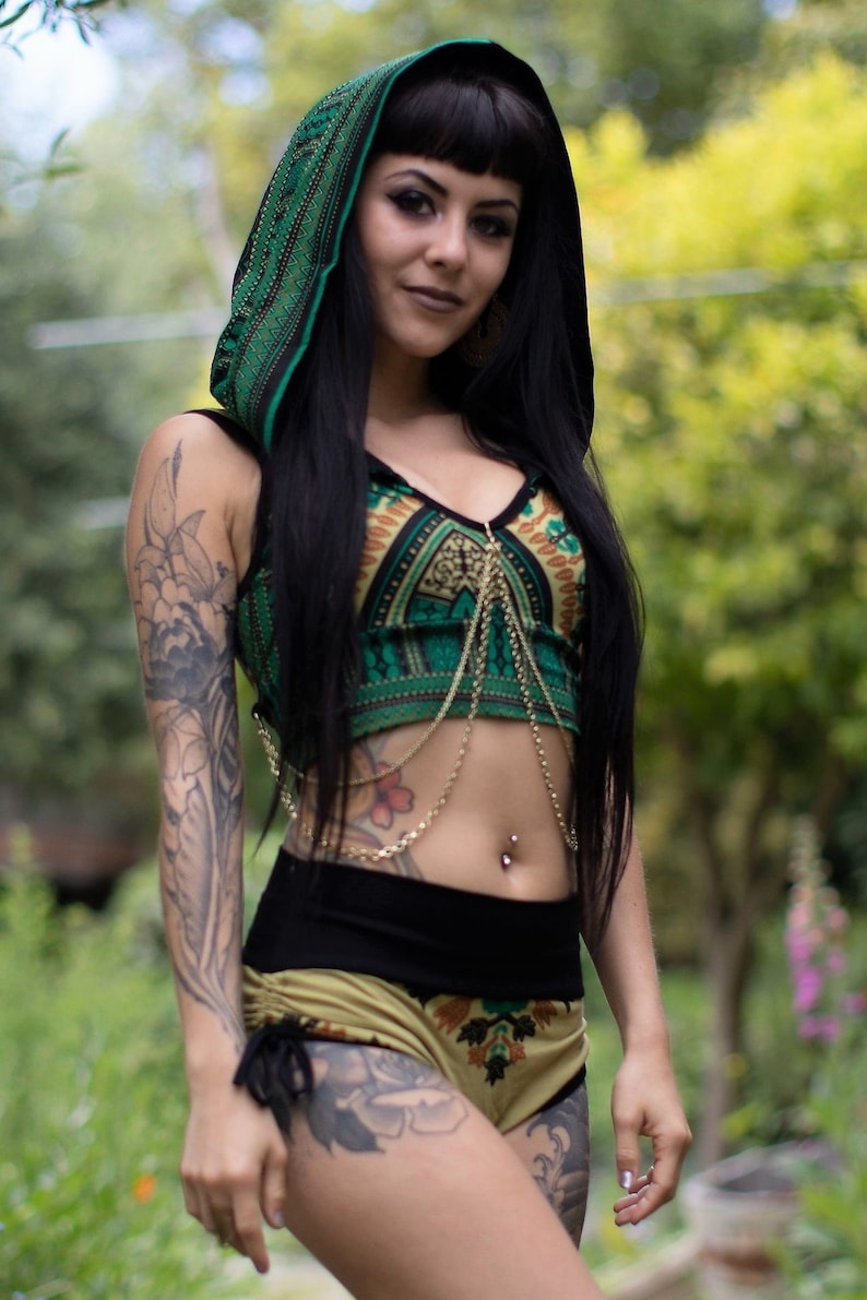 Puede incluir: Una mujer con un top con capucha de estampado verde y negro, un arn&eacute;s de cuerpo de cadena dorada y pantalones cortos a juego. Tiene el pelo negro y tatuajes en los brazos.