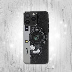 Puede incluir: Una funda para teléfono negra y plateada con un diseño de cámara vintage. La funda tiene una textura de piel sintética y una lente plateada con un borde negro.