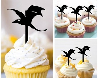 May include: Black silhouette dragon cupcake toppers. The dragon is flying with its wings spread out. The toppers are on top of cupcakes with white frosting and sprinkles.