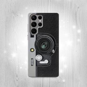 Puede incluir: Una funda de teléfono negra con un diseño de cámara vintage. La funda tiene un borde plateado y una textura de cuero negro. El objetivo de la cámara es visible en la parte posterior de la funda.