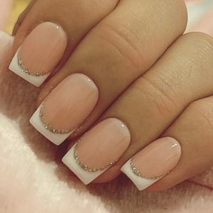 May include: Close-up of four fingernails with a white French manicure and a thin line of gold glitter along the edge of the white tip.