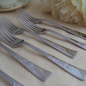 Könnte beinhalten: Ein Set aus sechs silbernen Gabeln mit verzierten Blumendesigns auf den Griffen. Die Gabeln sind in einer Reihe auf einer weißen Tischdecke angeordnet.