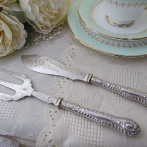 Puede incluir: Un juego de cuchillo y tenedor para pastel de plata con mangos adornados. El juego está sobre un mantel de encaje blanco.