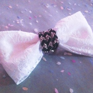 May include: A white lace bow with a black and silver center. The bow is made of delicate lace and has a classic bow shape. The center of the bow is a decorative piece with black and silver accents. The background is a soft, speckled pattern.
