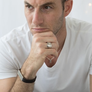 Puede incluir: Un hombre con una camiseta blanca, un brazalete de cuero marrón y un anillo de plata en el dedo. También lleva un collar de cadena de plata.