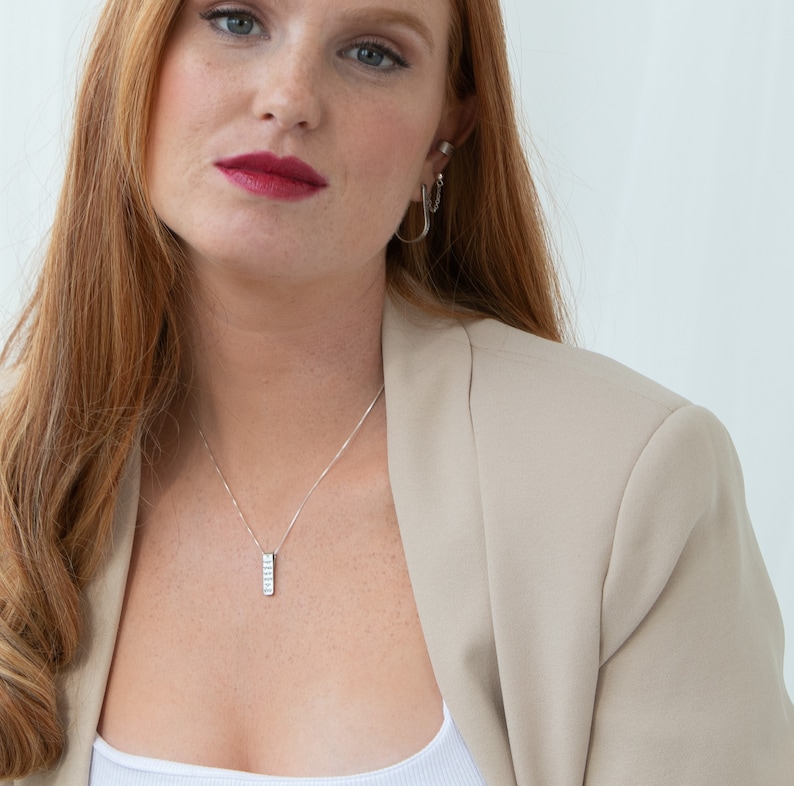 May include: A woman with long, red hair wearing a beige blazer and a silver necklace with a small, rectangular pendant.