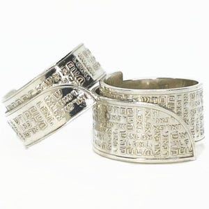 May include: Two silver rings with engraved Hebrew text. The rings have a curved design and are set against a white background. The rings are likely jewelry.
