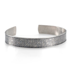 Puede incluir: Una pulsera de plata con un patrón texturizado de texto hebreo. La pulsera abierta tiene un diseño curvo y un acabado pulido. La palabra "KAD" está estampada en el interior de la pulsera.