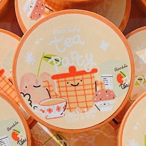 May include: A selection of circular stickers with an orange edge and a pale yellow backdrop. Each sticker showcases a cartoon tea party scene, with a teapot, teacup, and cupcake. The text "Bloom & Bake Tea Party" is displayed on each sticker.