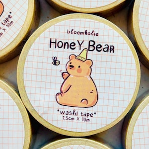 Op de afbeelding: Washi tape rollen met een cartoon honingbeer ontwerp. Elke rol heeft een lichtoranje beer met een gele hoed, met de tekst "Honey Bear" en "washi tape" gedrukt op een rasterachtergrond. De tape is 1,5 cm breed en 10 m lang.